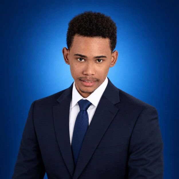 Professional head-and-shoulders portrait of a man in a navy suit and blue tie against a blue studio backdrop mis-lighting neutral expression