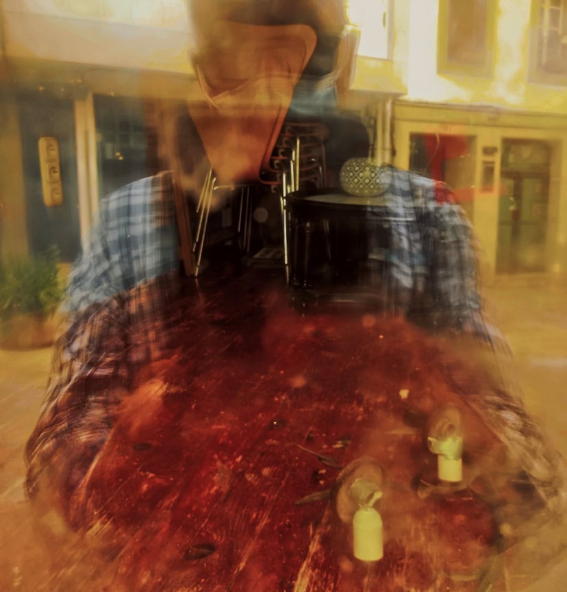 A double-exposure of a man in a blue plaid shirt reflected in glass, over a dark wooden table with a microphone stand visible in the background.