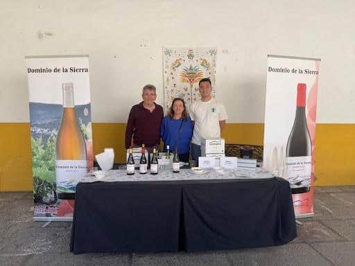 Three people stand behind a wine-tasting booth with bottles on the table and Dominio de la Sierra banners on either side.
