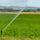 Center-pivot irrigation sprinkler watering a green field with a blue-roof barn in the background.