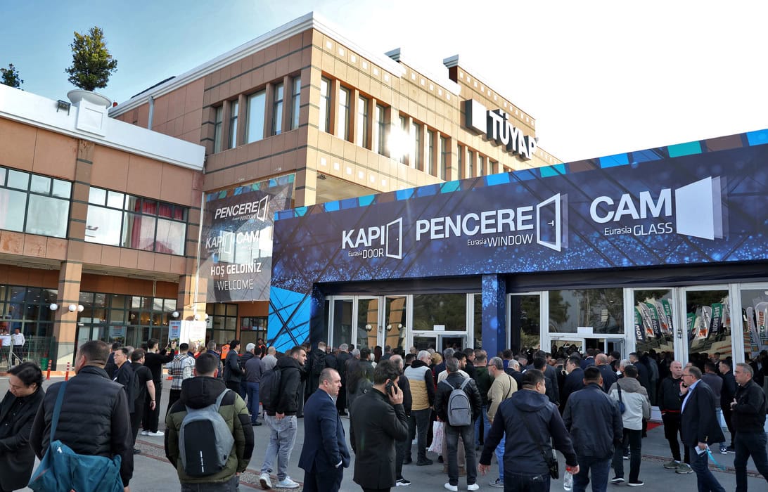 Crowd gathered outside a modern building with a large blue banner advertising window and glass brands, at an event entrance.