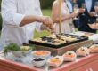 Chef grilling bite-size pastries on a hot griddle at an outdoor event, guests with drinks visible in the background.
