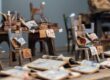 Miniature wooden chairs piled with colorful paper sketches and scrolls in a cluttered art studio.