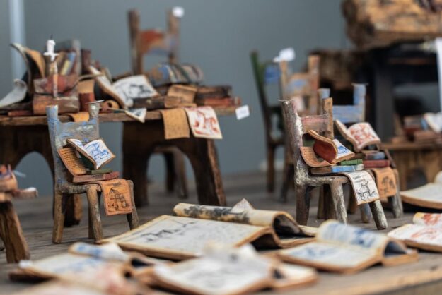 Miniature wooden chairs piled with colorful paper sketches and scrolls in a cluttered art studio.