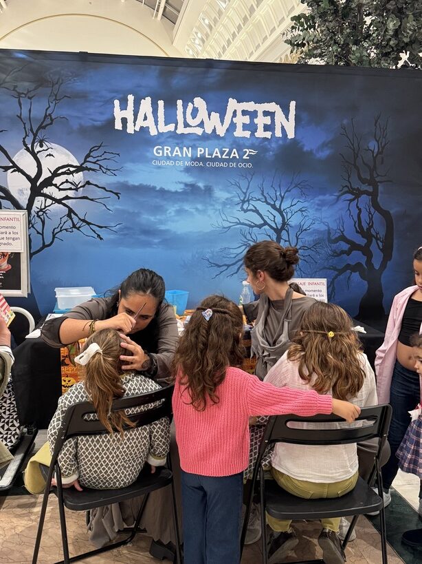 Group of kids getting Halloween makeup applied by adults at a mall event under a blue Halloween banner reading 'HALLOWEEN' and 'GRAN PLAZA 2°'.