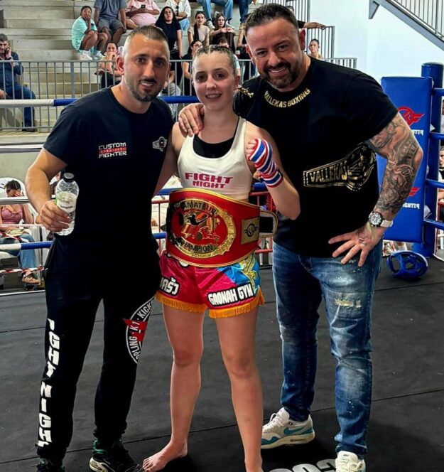 Young female boxer in a boxing ring posing with two men, wearing a championship belt and hand wraps, crowd behind her.