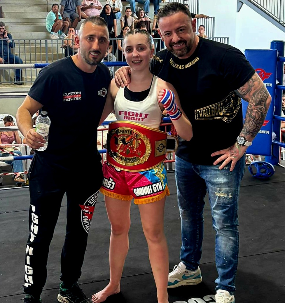 Young female boxer in a boxing ring posing with two men, wearing a championship belt and hand wraps, crowd behind her.