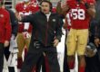 San Francisco 49ers coach on the sideline shouting commands with a headset, players in red and gold behind him.