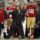 San Francisco 49ers coach on the sideline shouting commands with a headset, players in red and gold behind him.