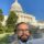 Selfie of a man in a suit with glasses in front of the U.S. Capitol dome against a blue sky.