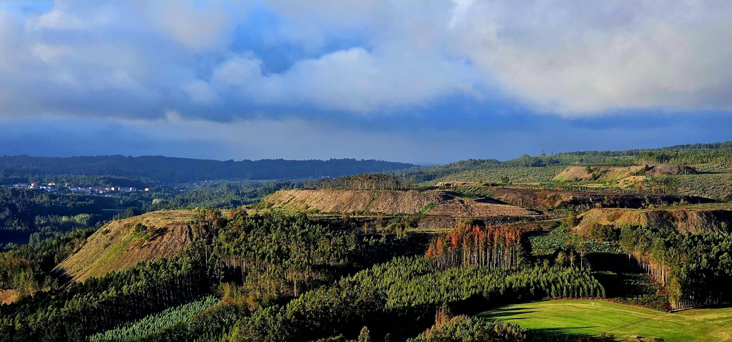 Wide view of rolling hills and patchwork fields under a cloudy blue sky, with a distant village along the horizon and dense forest in the foreground.