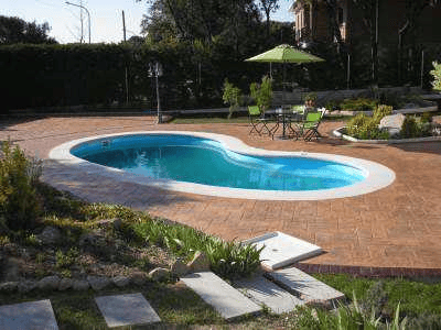 Kidney-shaped in-ground pool in a brick patio, with a green umbrella table set and surrounding garden.