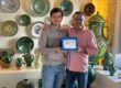 Two men pose in a ceramic shop, holding a blue framed award that reads 'Homo Faber'.
