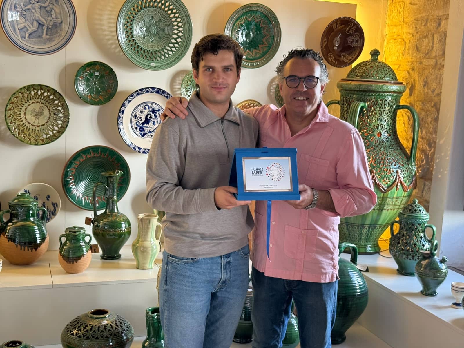 Two men pose in a ceramic shop, holding a blue framed award that reads 'Homo Faber'.