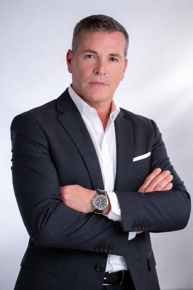 Professional man in a dark suit with white shirt and pocket square, arms crossed, looking at the camera.