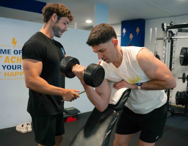Personal trainer spots a client performing a dumbbell bicep curl on a preacher bench in a gym.