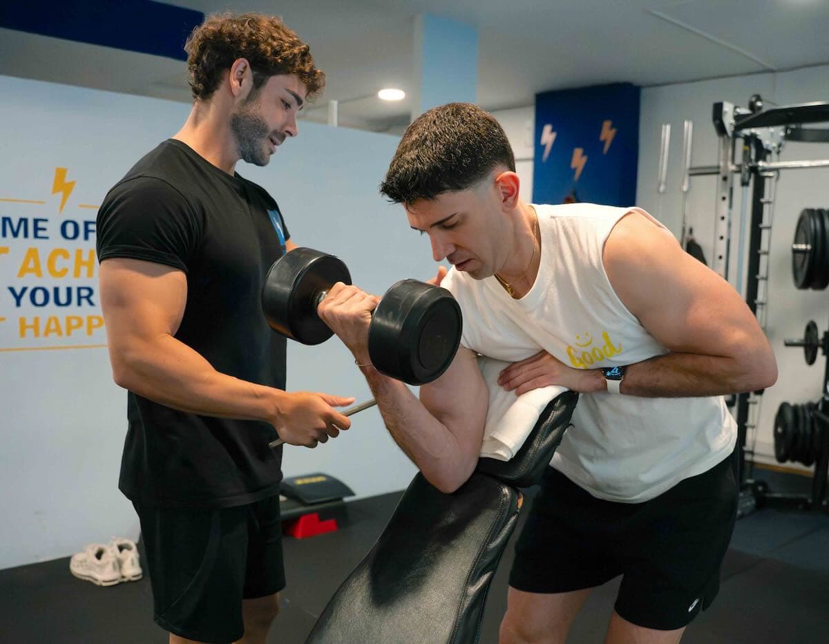 Personal trainer spots a client performing a dumbbell bicep curl on a preacher bench in a gym.