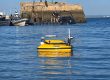 Yellow unmanned surface vehicle (USV) with sensor mast and antenna floats on blue harbor water, with a concrete breakwater and people on the pier in the background.