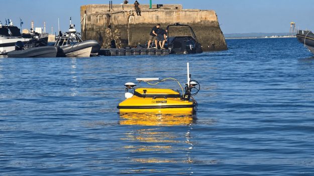 Yellow unmanned surface vehicle (USV) with sensor mast and antenna floats on blue harbor water, with a concrete breakwater and people on the pier in the background.