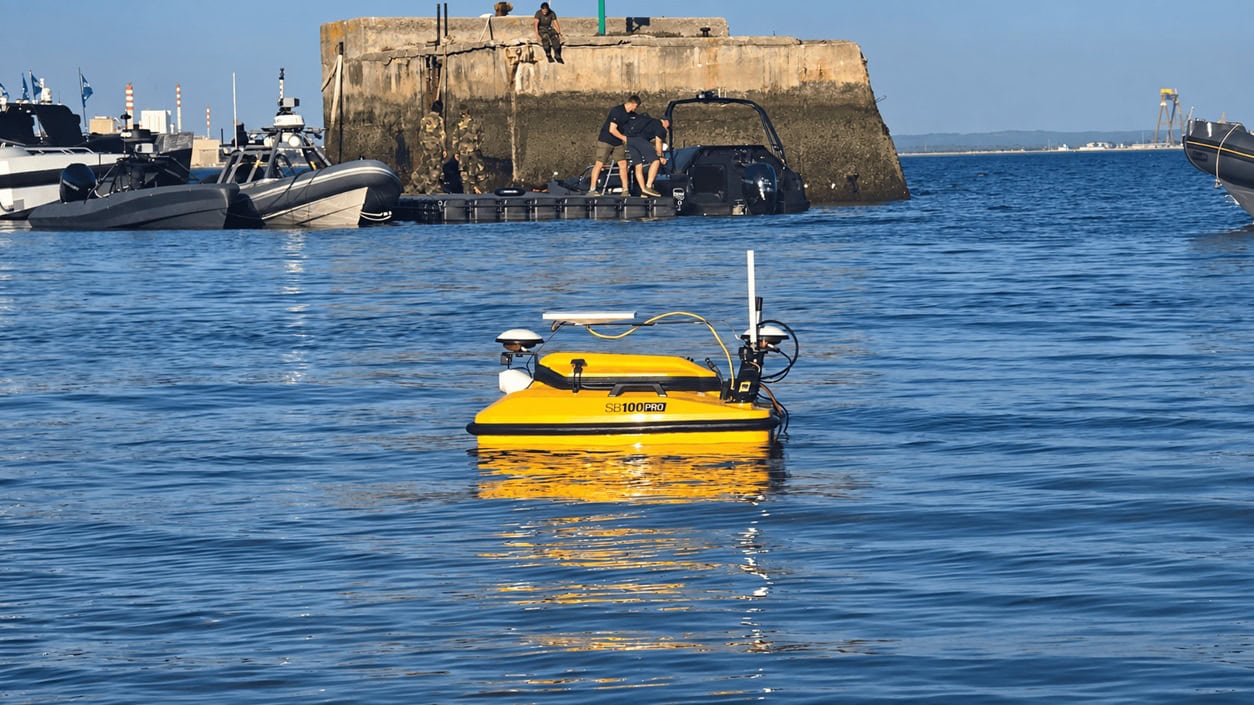 Yellow unmanned surface vehicle (USV) with sensor mast and antenna floats on blue harbor water, with a concrete breakwater and people on the pier in the background.