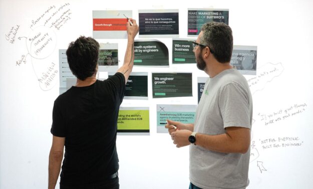 Two people stand with their backs to the camera, writing on a wall filled with posters and notes.
