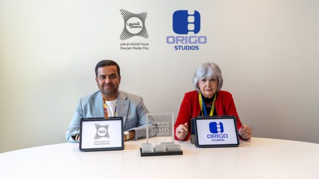 Two people sit at a round white table with tablets showing Shams and ORIGO Studios logos, against a light wall with the same logos behind them, a man in a gray blazer and a woman in red.