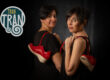 Two women stand back-to-back in black outfits, each holding red sneakers, against a dark backdrop with a teal and white TRAN logo to the left.