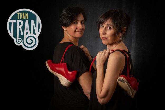 Two women stand back-to-back in black outfits, each holding red sneakers, against a dark backdrop with a teal and white TRAN logo to the left.