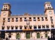 Stone town hall with two towers and a row of arched windows, displaying multiple Spanish flags along the façade; people walk in the plaza.