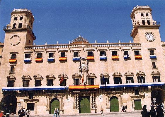 Stone town hall with two towers and a row of arched windows, displaying multiple Spanish flags along the façade; people walk in the plaza.