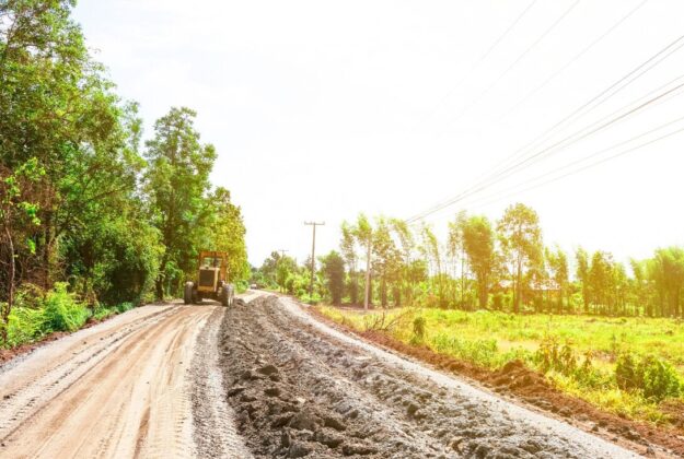 Dusty rural road under construction with a yellow bulldozer on the left and trees lining the road under a bright sky, power lines overhead.