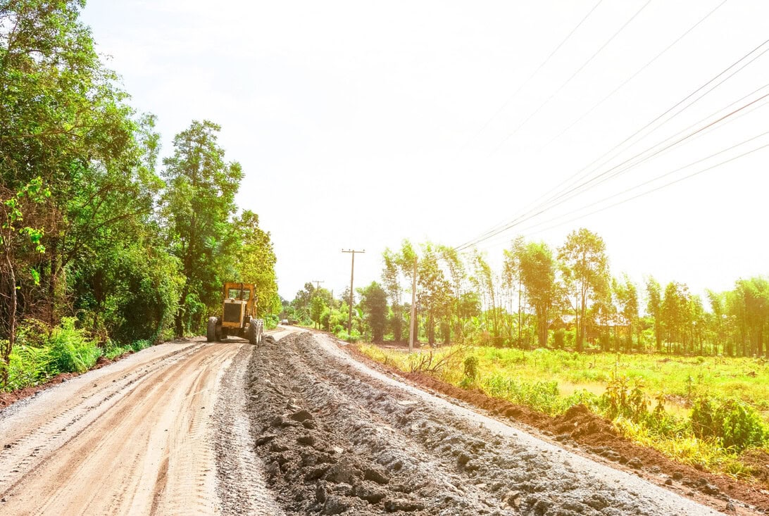 Dusty rural road under construction with a yellow bulldozer on the left and trees lining the road under a bright sky, power lines overhead.