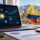 Laptop screen shows a cryptocurrency analytics dashboard with world map connections and charts; a Colombia-shaped map in its flag colors (yellow, blue, red) sits nearby with Bitcoin coins on the desk.