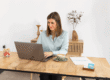 Woman sits at a wooden table typing on a laptop, with a coffee cup and papers nearby in a bright, minimal office.