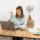 Woman sits at a wooden table typing on a laptop, with a coffee cup and papers nearby in a bright, minimal office.