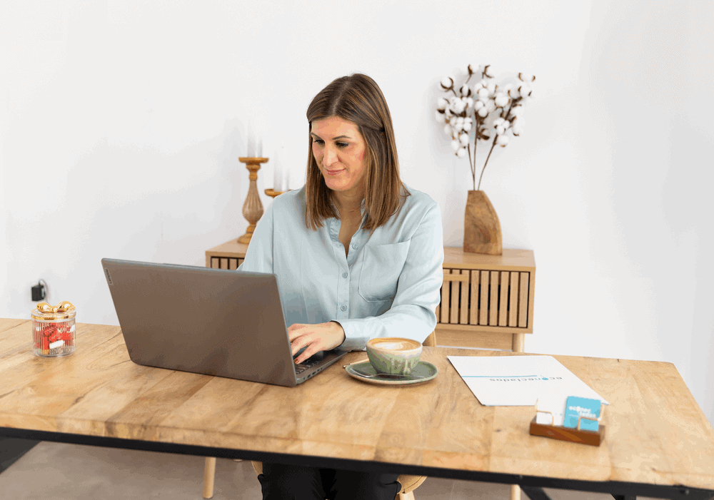 Woman sits at a wooden table typing on a laptop, with a coffee cup and papers nearby in a bright, minimal office.