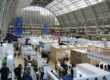 Interior of a large glass-roofed exhibition hall filled with white booths and attendees browsing displays at a trade fair.