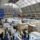 Interior of a large glass-roofed exhibition hall filled with white booths and attendees browsing displays at a trade fair.