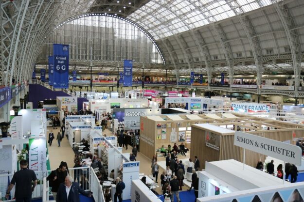 Interior of a large glass-roofed exhibition hall filled with white booths and attendees browsing displays at a trade fair.