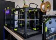 Black 3D printer frame with neon-green clamps on a workbench and spool holders with filament in a lab setting