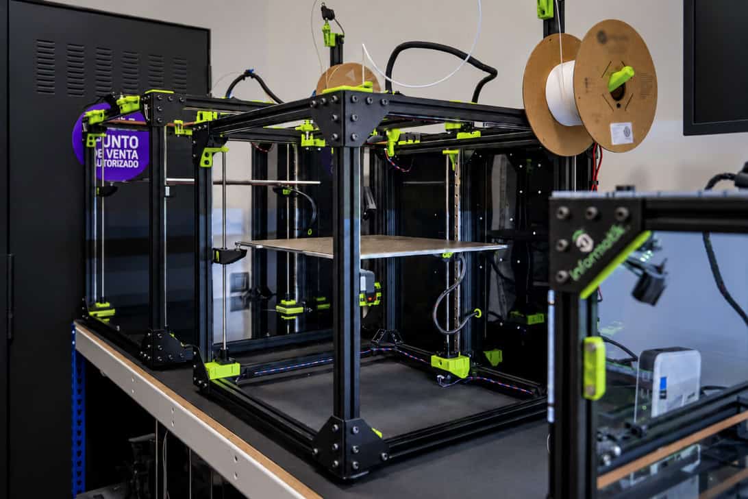 Black 3D printer frame with neon-green clamps on a workbench and spool holders with filament in a lab setting