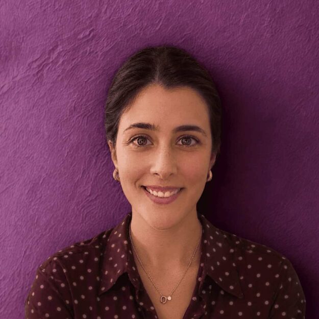 Smiling woman with dark hair against a purple wall, wearing a polka-dot shirt and necklace.