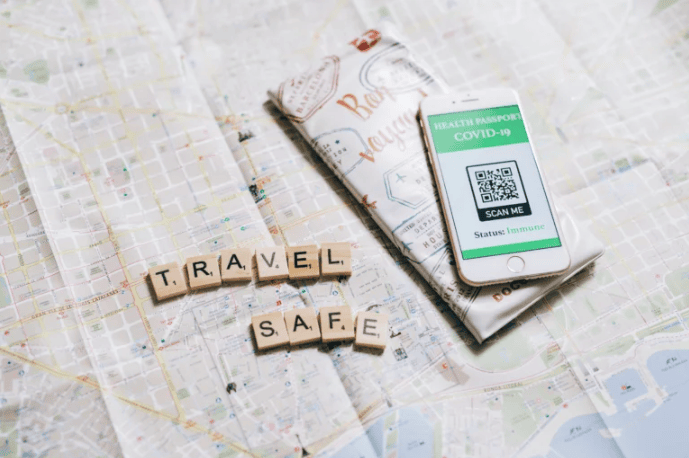 Map with Scrabble tiles spelling 'TRAVEL SAFE' beside a wallet and a phone displaying a COVID-19 health passport: 'SCAN ME' QR code and immune status.