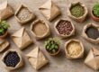 Top-down view of brown paper bags filled with assorted seeds and grains on a wooden table, with two small potted plants nearby.