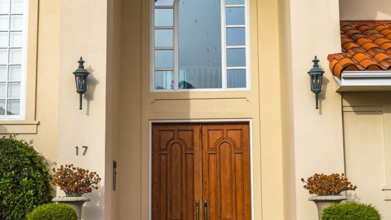 Arched stucco entry of a tidy Southwest home with double wooden doors and trimmed shrubs