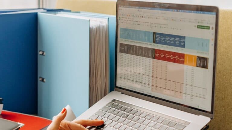 Hands typing on a laptop displaying a color coded spreadsheet beside office binders and a bright window