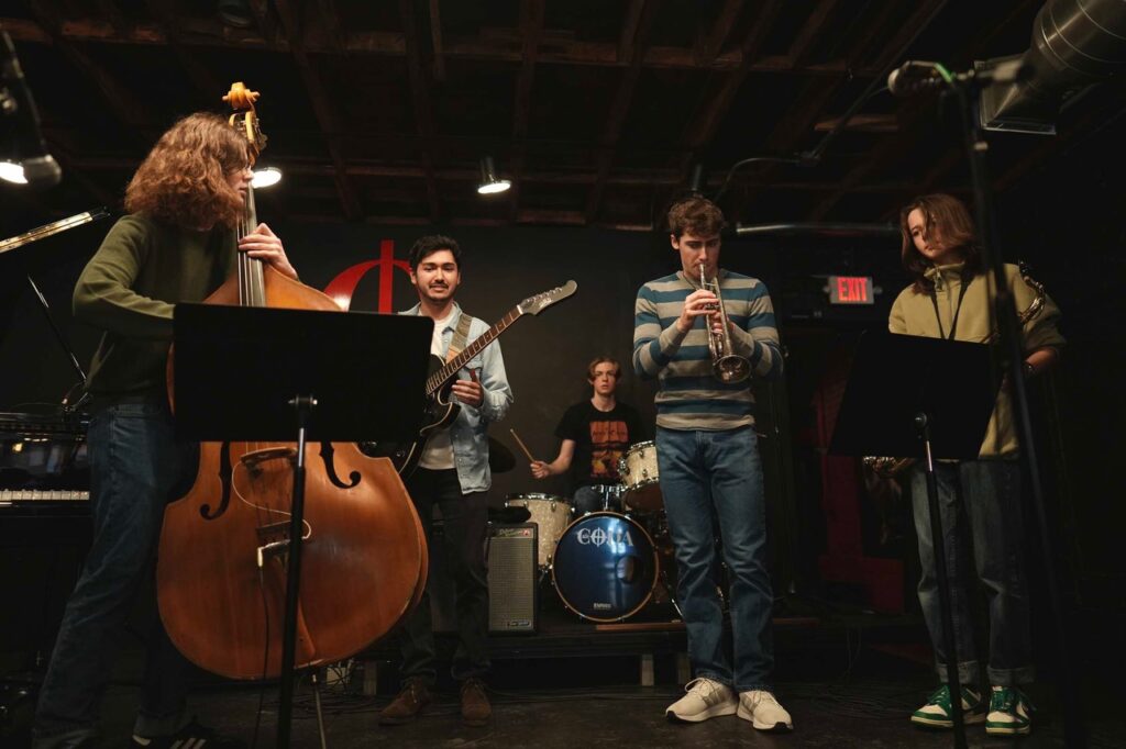 A student jazz combo performs on a dimly lit stage with upright bass, guitar, drums, and horns