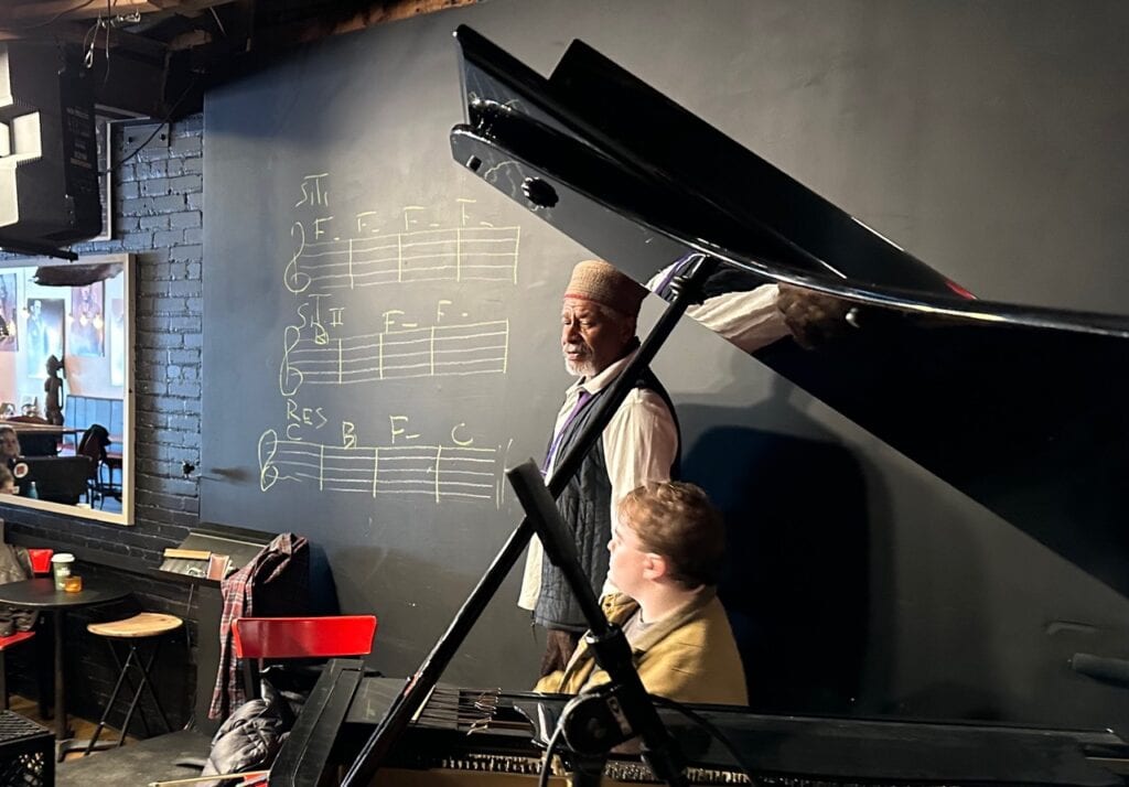 An instructor plays a grand piano beside a young child, with musical notation on the chalkboard behind them