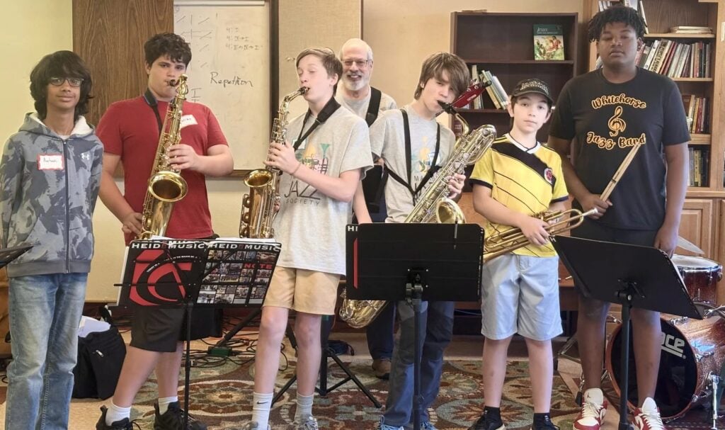 Young musicians with saxophones, a baritone horn and drumsticks stand together during an instrument-specific session at iGnitE jAzz!, with an instructor behind them