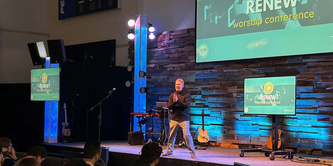 Speaker at NLW International worship conference on stage with screens and musical instruments.
